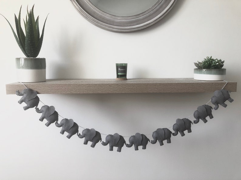 May include: A gray felt elephant garland strung on a white string, hanging below a wooden shelf. The garland is displayed with two potted plants and a small candle. The garland is a decorative item.