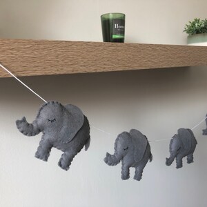 May include: A gray felt elephant garland strung on a white cord. Each elephant has closed eyes and is in a walking pose. The garland is displayed against a neutral background, suggesting a nursery or children's room decoration.