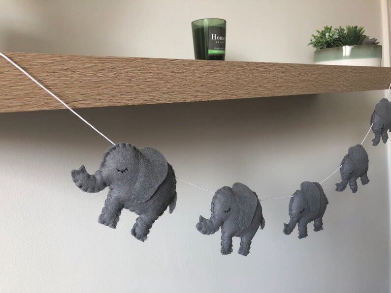 May include: A gray felt elephant garland strung on a white cord. Each elephant has closed eyes and is in a walking pose. The garland is displayed against a neutral background, suggesting a nursery or children's room decoration.