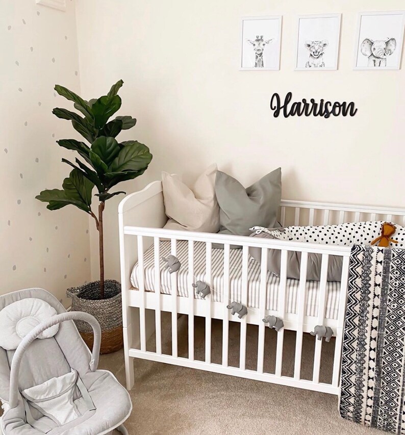 May include: A white crib with gray and white striped bedding, elephant-shaped decorations, and a black and white patterned blanket. The wall features framed animal prints and a black name sign that reads "Harrison". A gray baby bouncer and a potted plant are also in the room.