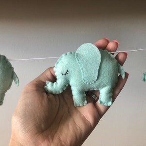 May include: A garland of light teal felt elephants strung on a white cord. Each elephant has a stitched outline, a small eye, and a trunk. The garland is held in a hand.