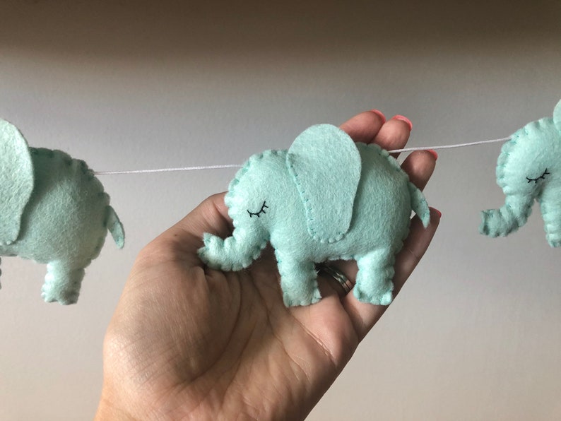 May include: A garland of light teal felt elephants strung on a white cord. Each elephant has a stitched outline, a small eye, and a trunk. The garland is held in a hand.