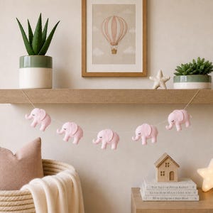 May include: A nursery decor scene featuring a wooden shelf with potted plants, a framed hot air balloon print, and a string of pink elephant ornaments. A woven basket with a pillow and blanket is in the foreground.