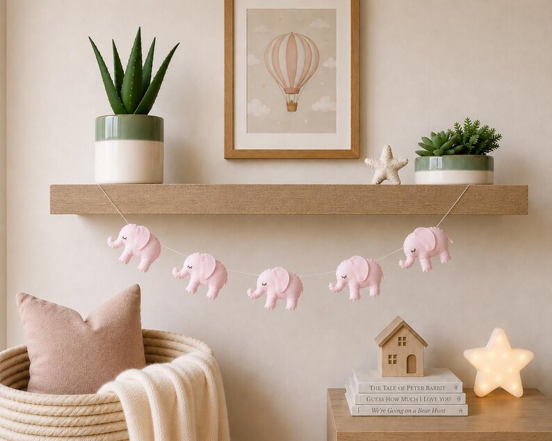 May include: A nursery decor scene featuring a wooden shelf with potted plants, a framed hot air balloon print, and a string of pink elephant ornaments. A woven basket with a pillow and blanket is in the foreground.