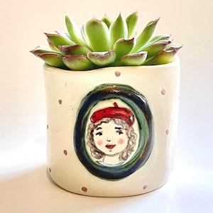 May include: A ceramic plant pot with a green succulent. The off-white pot features a hand-painted portrait of a woman wearing a red beret. The pot has pink polka dots and a dark blue and green oval frame around the portrait.