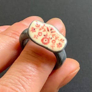 May include: A ring featuring a dark grey band and a rectangular, slightly curved top piece. The top surface is cream-colored with a detailed, repeating floral pattern in shades of red and orange, creating a distinct contrast against the dark band.