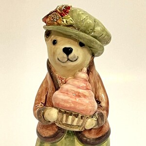 May include: A ceramic bear figurine wearing a green hat and brown coat, holding a pink cupcake. The bear has a friendly expression and is decorated with detailed paintwork. The figurine is a decorative item.