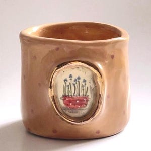 May include: A ceramic container with a light brown exterior and small pink polka dots. An oval-shaped inset features a painting of blue flowers in a red dotted pot. The container has a smooth, rounded shape.