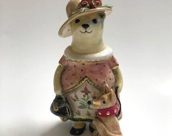 May include: A ceramic figurine of a bear wearing a cream-colored hat with red roses, a pink polka-dot dress with a floral apron, and black shoes. The bear holds a black handbag and stands next to a small tan dog wearing a bright pink collar.