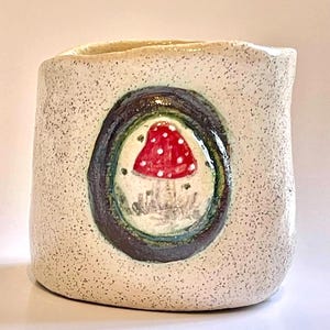 May include: A speckled ceramic mug featuring a hand-painted mushroom design. The mushroom has a red cap with white spots, set within a dark brown and green oval. The mug is off-white with a textured surface.