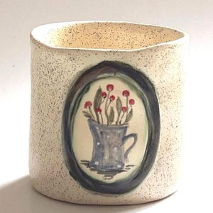 May include: A ceramic vase with a speckled beige exterior. An oval painting on the front depicts a blue pitcher with white polka dots, holding red-topped flowers. The painting is framed in a dark oval.