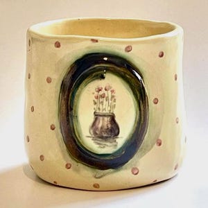 May include: A handmade ceramic vase with a cream-colored base, adorned with small pink polka dots. The vase features a painted oval with a floral still life. The oval is framed by a dark blue and green border.