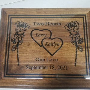 Puede incluir: Una placa de madera con un borde grabado en negro y dos rosas a cada lado de dos corazones entrelazados. Los corazones están grabados con los nombres "Larry" y "Kaitlyn". La placa también incluye el texto "Two Hearts, One Love, September 18, 2021".