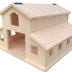 Puede incluir: Una casa de muñecas de madera clara con forma de granero, con una estructura principal de dos pisos y una extensión de un piso. El techo tiene una sección con bisagras. El diseño incluye múltiples ventanas rectangulares y una gran abertura en la planta baja.