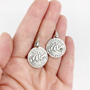 May include: A pair of silver dangle earrings featuring a vintage half penny coin with a ship design. The coin has the text "HALF PENNY" and "1961" on it.