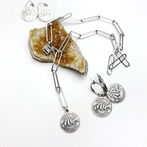 May include: A silver necklace with a silver charm of a ship and a silver chain. The necklace is on a piece of yellow rock. There are also two silver earrings with ship charms.