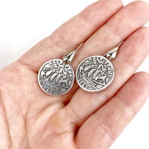 May include: A pair of silver dangle earrings featuring a round coin design with a ship and the text "Half Penny" on each earring.