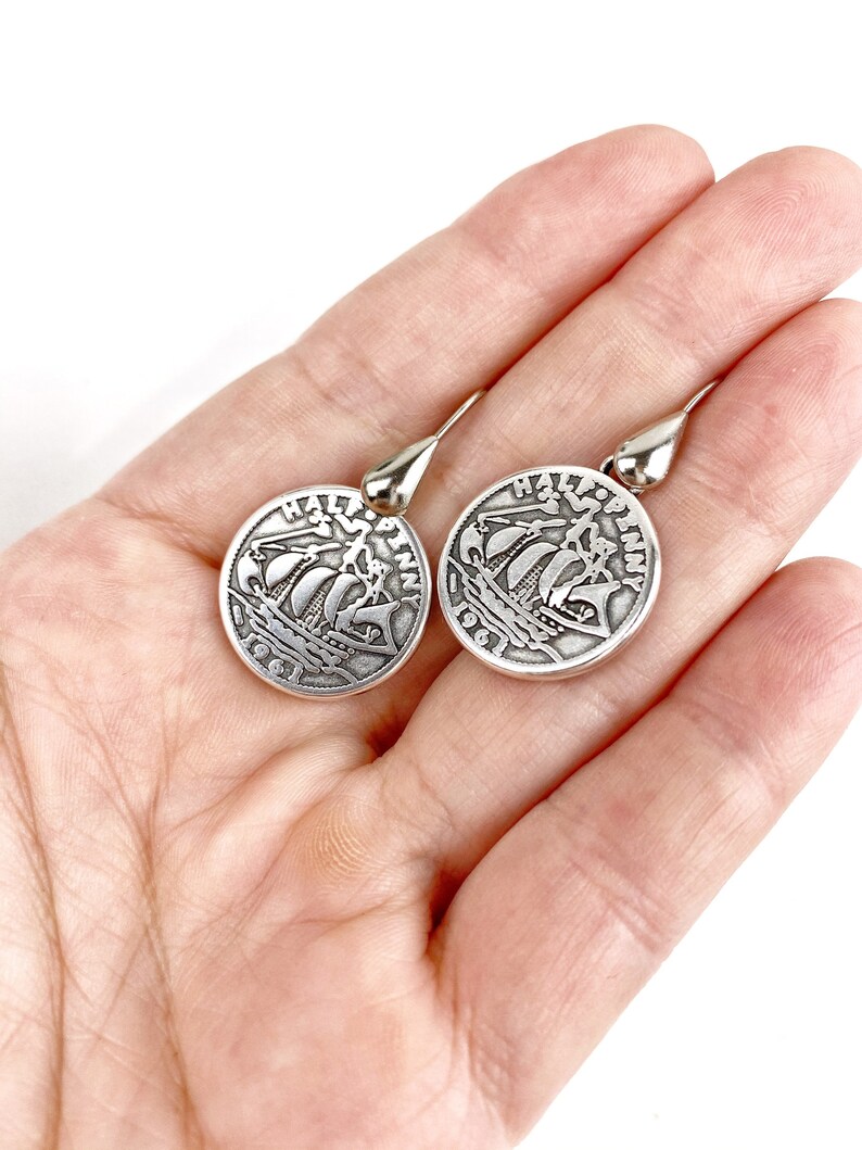 May include: A pair of silver dangle earrings featuring a round coin design with a ship and the text "Half Penny" on each earring.