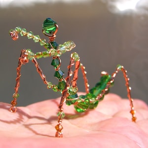 以下が含まれることがあります： 緑と透明なクリスタルビーズで飾られた、手作りの銅線で作られた昆虫の彫刻。昆虫は6本の脚、分割された体、そして伸びた腕を持ち、すべて複雑なディテールで作成されています。ニュートラルな背景に表示されています。