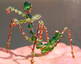 May include: A handcrafted copper wire sculpture of an insect, adorned with green and clear crystal beads. The insect has six legs, a segmented body, and outstretched arms, all crafted with intricate detail. The sculpture is displayed on a neutral background.