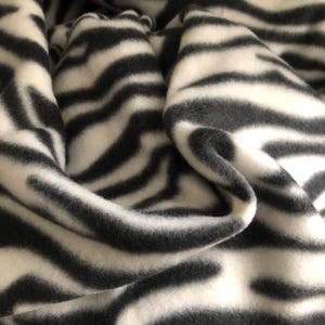 K&ouml;nnte beinhalten: Nahaufnahme einer gefalteten Decke mit Zebrastreifenmuster. Die Decke hat abwechselnd schwarze und wei&szlig;e Streifen, die ein auff&auml;lliges Design erzeugen. Der Stoff wirkt weich und gem&uuml;tlich, ideal f&uuml;r die Inneneinrichtung.