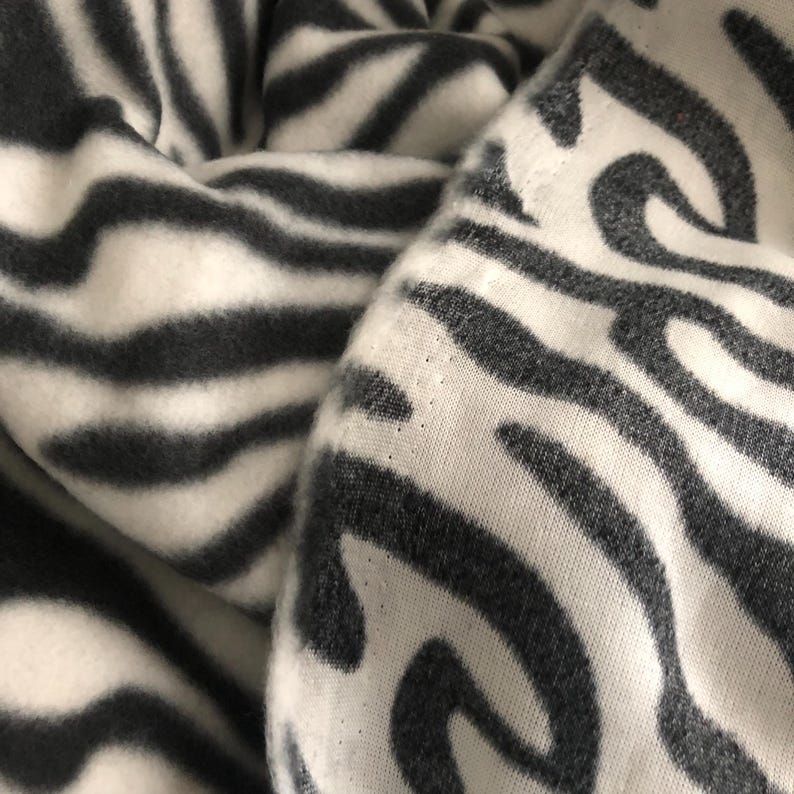 K&ouml;nnte beinhalten: Nahaufnahme eines Stoffes mit Zebrastreifenmuster. Der Stoff ist wei&szlig; mit dunkelgrauen Streifen, was einen kr&auml;ftigen Kontrast erzeugt. Das Material wirkt weich und m&ouml;glicherweise gestrickt oder gewebt, mit sichtbarer Textur und Falten.