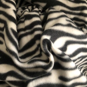 K&ouml;nnte beinhalten: Nahaufnahme eines gefalteten Stoffes mit Zebrastreifenmuster. Der Stoff hat abwechselnd schwarze und wei&szlig;e Streifen, die ein auff&auml;lliges Design ergeben. Das Material scheint weich und bequem zu sein, ideal f&uuml;r Decken.