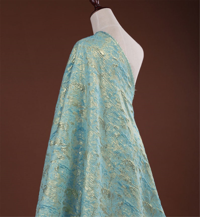 May include: A draped fabric in a light blue hue with a textured, metallic gold pattern. The fabric is displayed on a mannequin, showcasing its drape and texture. The material has a shimmering effect, suitable for apparel or decorative projects.