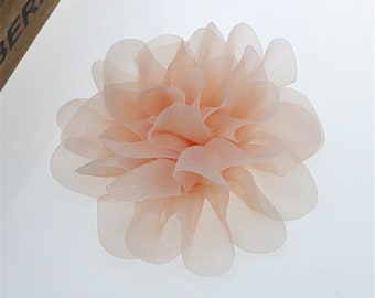 May include: A delicate peach-colored fabric flower with a layered, ruffled design. The flower is made of soft, sheer material and has a delicate, feminine look.