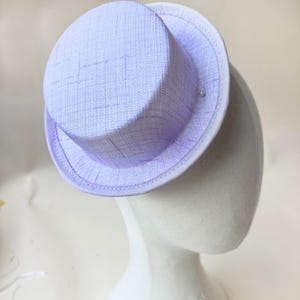 May include: A light lavender mini top hat featuring a textured, woven fabric finish and a crisp white trim along the edge of the brim. The hat has a flat crown and a short, structured brim, presenting a clean, uniform appearance throughout.