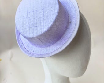 Puede incluir: Un mini sombrero de copa de color lavanda claro con un acabado de tela tejida texturizada y un borde blanco nítido a lo largo del ala. El sombrero tiene una corona plana y un ala corta y estructurada, presentando un aspecto limpio y uniforme.