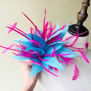 Könnte beinhalten: Ein auffälliger Fascinator mit leuchtend blauen und pinkfarbenen Federn. Die Federn sind in einem geschichteten, blütenartigen Design angeordnet, mit langen, schlanken Federn, die sich nach außen erstrecken. Ein Accessoire für besondere Anlässe.