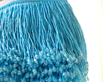 Pode incluir: Um close-up de uma guarnição de franja de contas azul claro. A franja é feita de pequenas contas redondas que são enfiadas em um fio fino. As contas são iridescentes e refletem a luz, criando um efeito brilhante.
