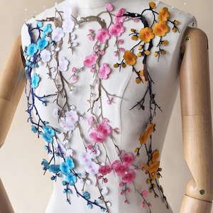 Puede incluir: Una aplicación bordada que presenta ramas en cascada con flores en tonos azul, blanco, rosa y amarillo. El diseño floral está dispuesto sobre un torso de maniquí blanco, mostrando costuras intrincadas y detalles delicados en los pétalos.