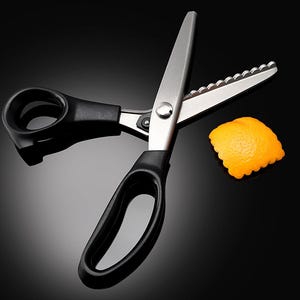 May include: Pinking shears with silver metal blades and black plastic handles. The blades feature a serrated edge designed for cutting fabric in a zigzag pattern. A small, square piece of orange fabric with a scalloped edge sits nearby.