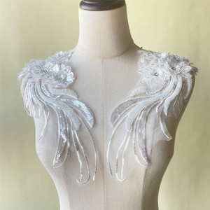 May include: Two white lace appliques with silver sequins and beads. The appliques are shaped like a wing and have a floral design at the top.