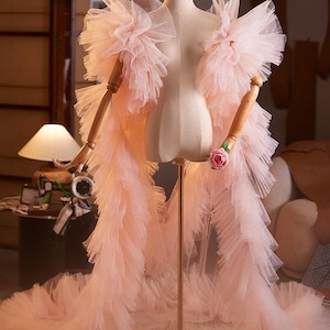 May include: A pale pink tulle garment featuring voluminous, layered ruffles that extend from the shoulders down to the floor. The piece is displayed on a mannequin with articulated wooden arms, with one hand holding a single pink rose.