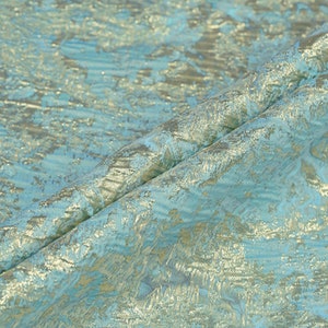 May include: Close-up of a folded fabric with a textured, shimmering surface. The fabric features a blend of light blue and gold colors, creating a luxurious and elegant appearance. The material has a subtle, abstract pattern.