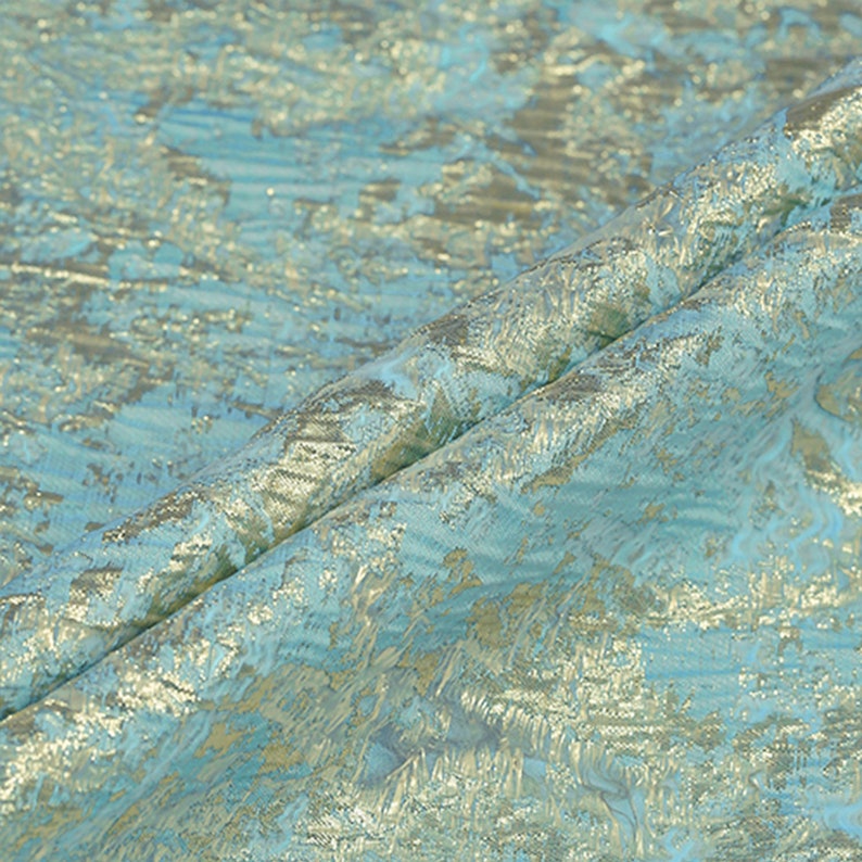 May include: Close-up of a folded fabric with a textured, shimmering surface. The fabric features a blend of light blue and gold colors, creating a luxurious and elegant appearance. The material has a subtle, abstract pattern.