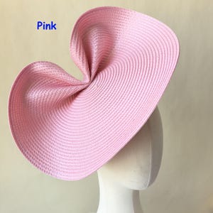 May include: A large, pink, woven hat with a unique, folded design. The hat is displayed on a mannequin head. The word "Pink" is written in blue in the upper left corner. The hat's brim has a curved shape.