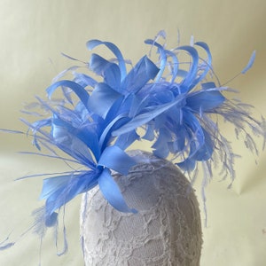 Puede incluir: Un tocado de plumas azul claro con un gran lazo y plumas largas y flotantes. El tocado está en una cabeza de maniquí de encaje blanco.