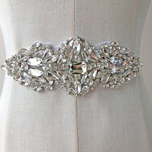 May include: A crystal embellished bridal belt with a delicate, wavy design. The belt is made of clear crystals of various sizes and shapes, sewn onto a sheer fabric base.