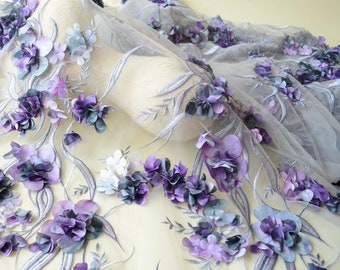 May include: A sheer fabric with 3D floral embellishments. The flowers are in shades of purple, blue, and white, with silver embroidered stems and leaves. The fabric is draped over a mannequin.