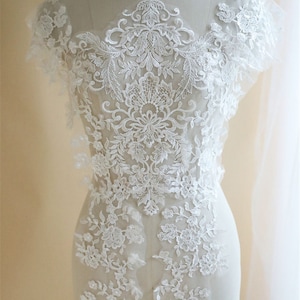 Op de afbeelding: Witte kanten applicatie met bloemen- en siermotieven. De delicate stof wordt op een mannequin getoond, waarbij de ingewikkelde details van het borduurwerk worden getoond. Deze versiering voor bruids- of formele kleding heeft een verscheidenheid aan bloempatronen.