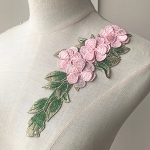 Op de afbeelding: Roze bloemenapplicatie met groene bladeren en een parel. Deze delicate borduurwerk is perfect om kleding, accessoires of woonaccessoires een vleugje elegantie te geven.