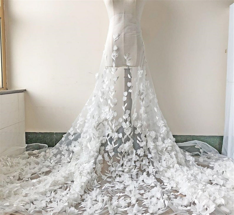 May include: White floral lace fabric with 3D flowers. The lace is delicate and intricate, with a soft, flowing texture. The flowers are white and have a realistic appearance.