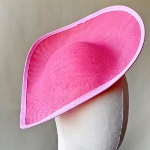 Puede incluir: Un sombrero rosa brillante con un ala curva y una copa redondeada. El sombrero está hecho de un material similar a una malla y está bordeado con un borde rosa más claro. El sombrero se exhibe en una cabeza de maniquí.