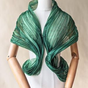 May include: A sheer emerald green shawl featuring dense vertical pleats throughout the fabric. The garment is draped around the neck and shoulders, creating a voluminous, layered effect with a translucent texture and a deep, vibrant green color.