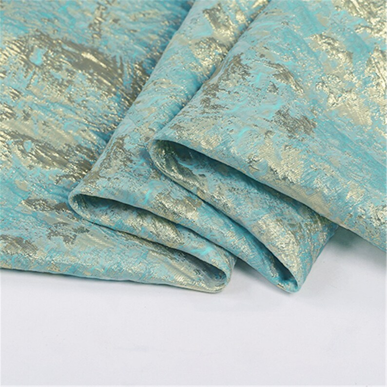 May include: Close-up of folded fabric with a textured pattern. The fabric is a light teal color with gold and silver metallic accents. The folds create shadows and depth, highlighting the intricate design. The fabric appears to be a luxurious material, possibly for apparel or home decor.