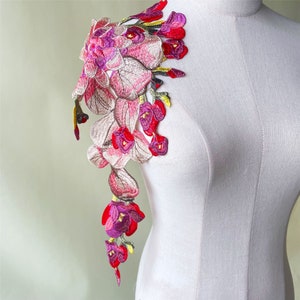 May include: A pink, purple and green embroidered floral applique. The applique features a cluster of flowers with delicate details.